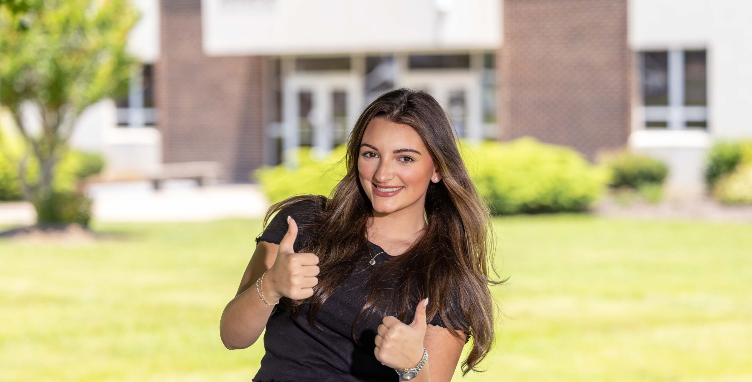 Female student holding thumbs up, sitting outside of Polkton campus grouind.
