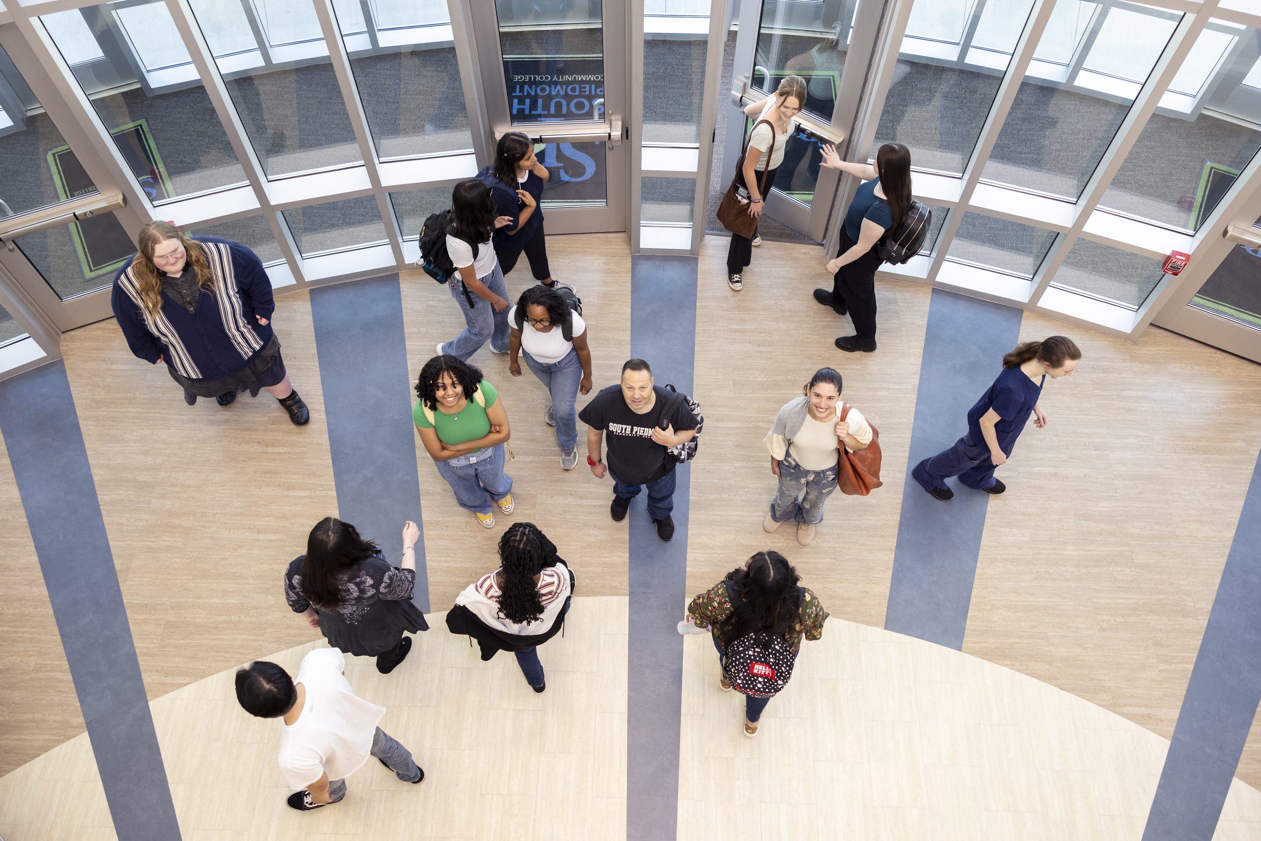 A shot from above, looking down at students walking in and out of the main entrance of the OCH Main building. In the center, three students pause and look up at the camera, smiling amid the flow of activity.