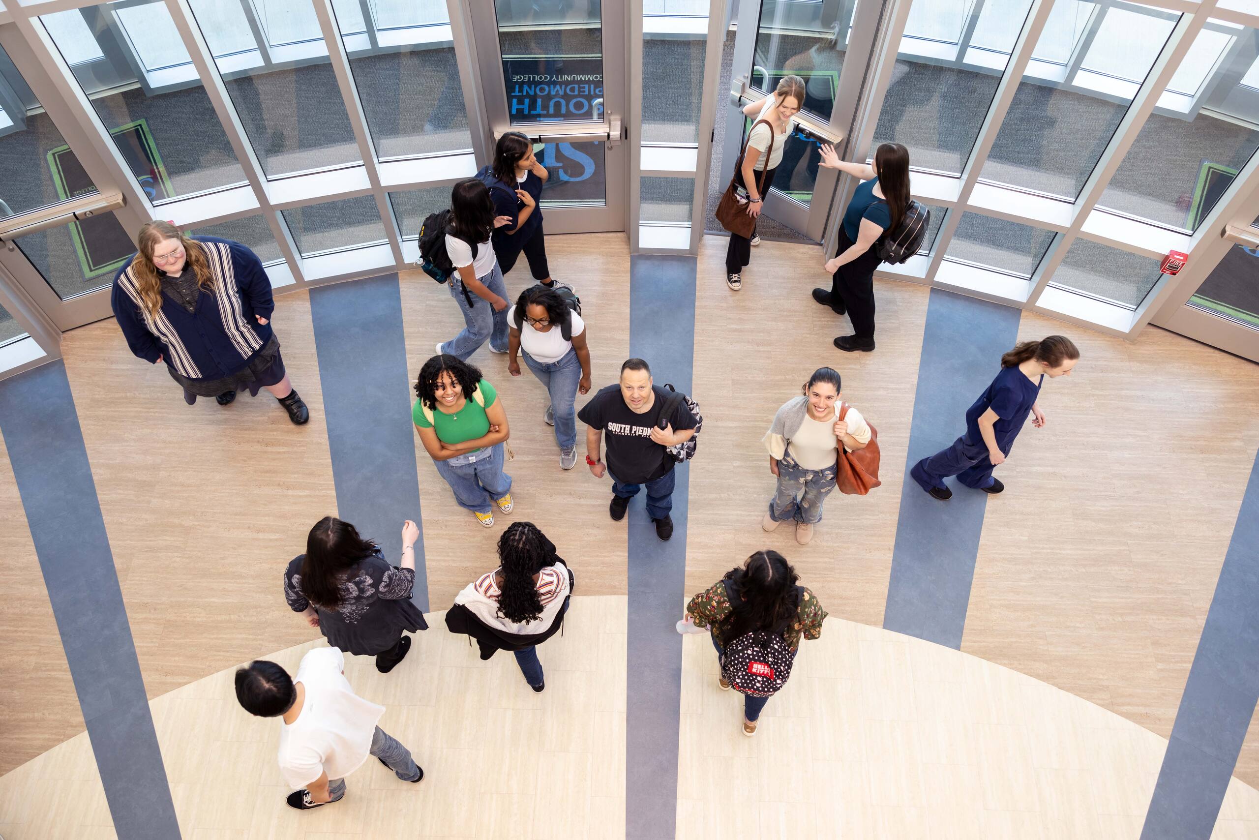 A shot from above, looking down at students walking in and out of the main entrance of the OCH Main building. In the center, three students pause and look up at the camera, smiling amid the flow of activity.
