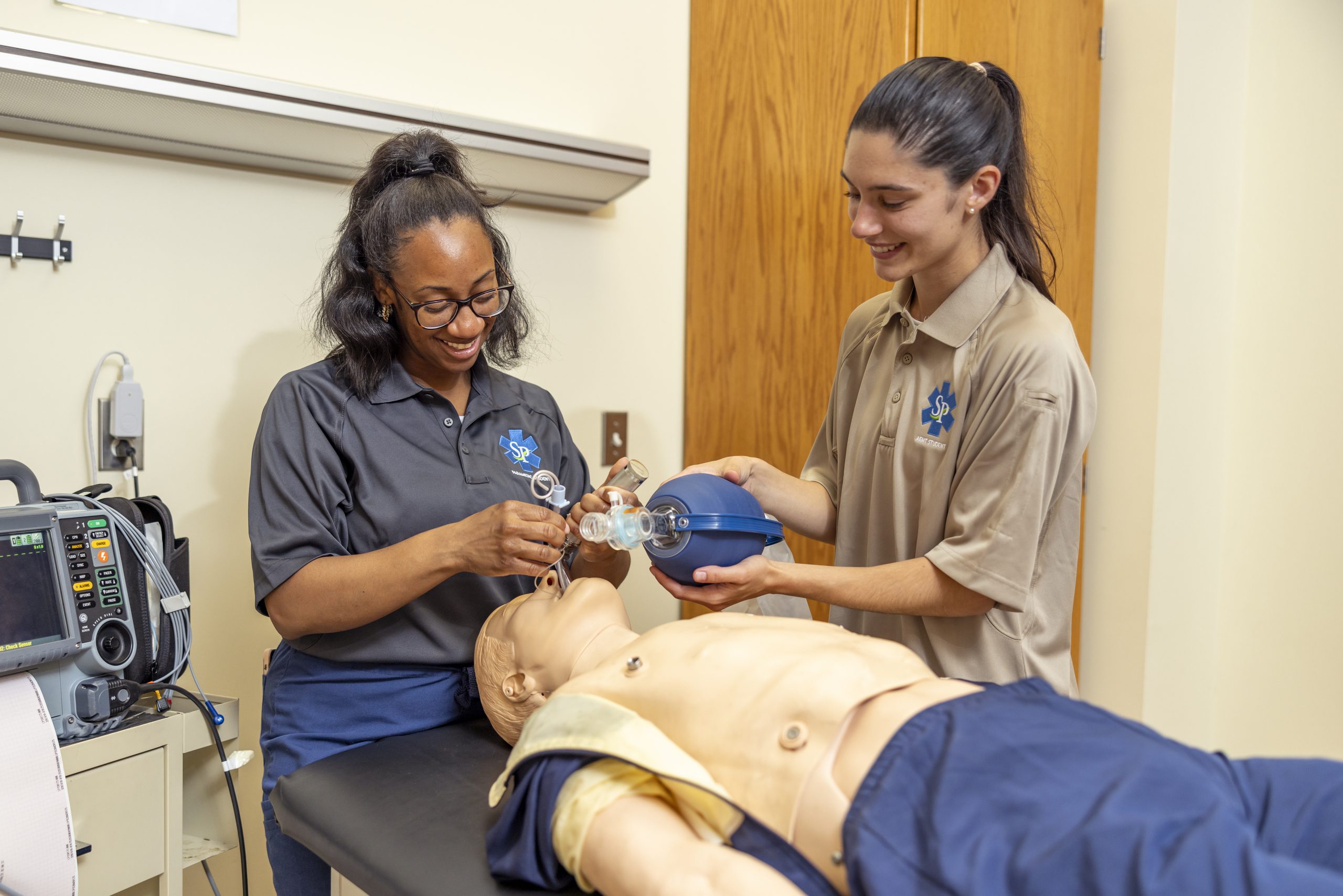 Two female students practicing CPR by using a manual resuscitator to pump air into a mannequin during a medical training session.