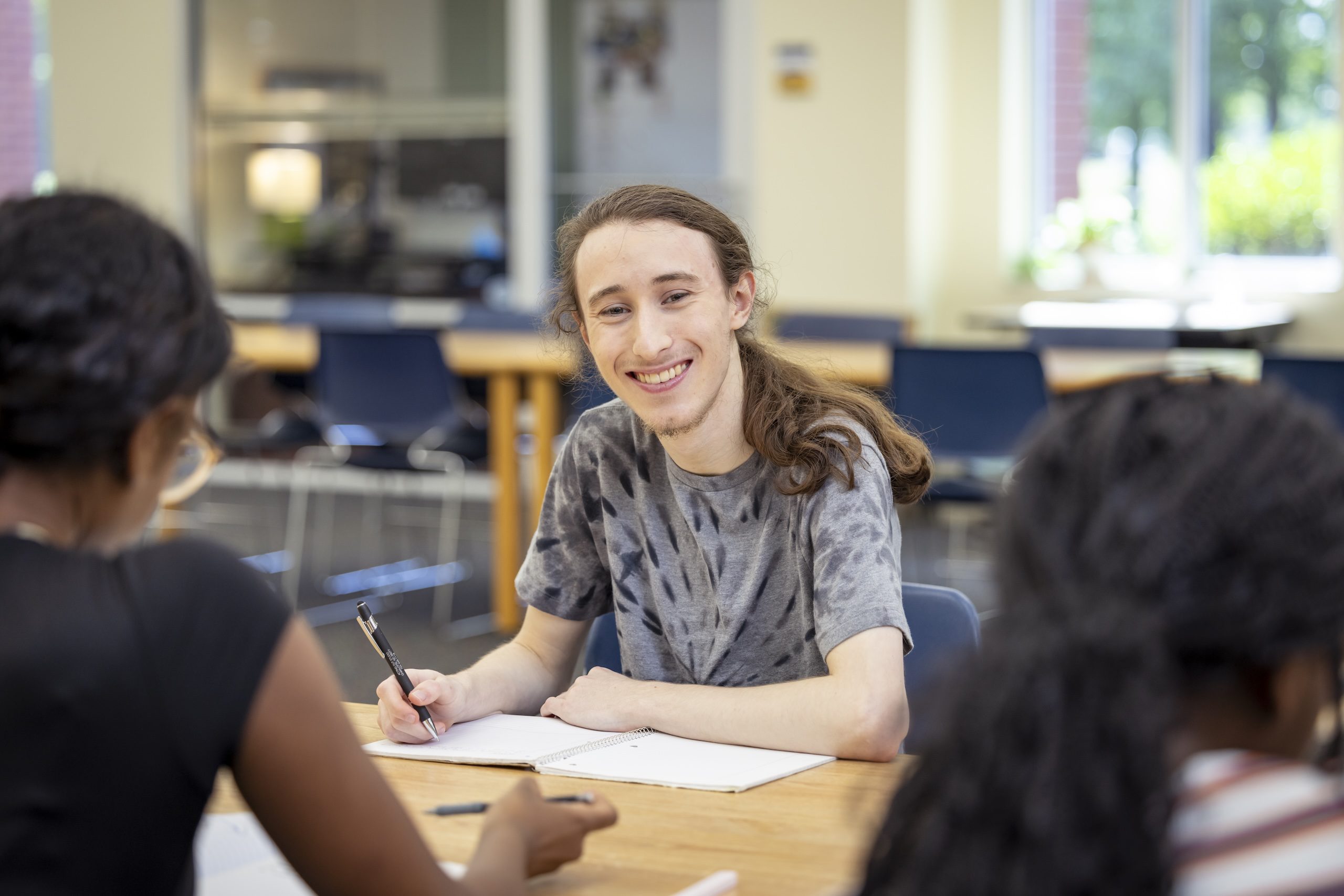Male student sitting inside the library, smiling at the camera.