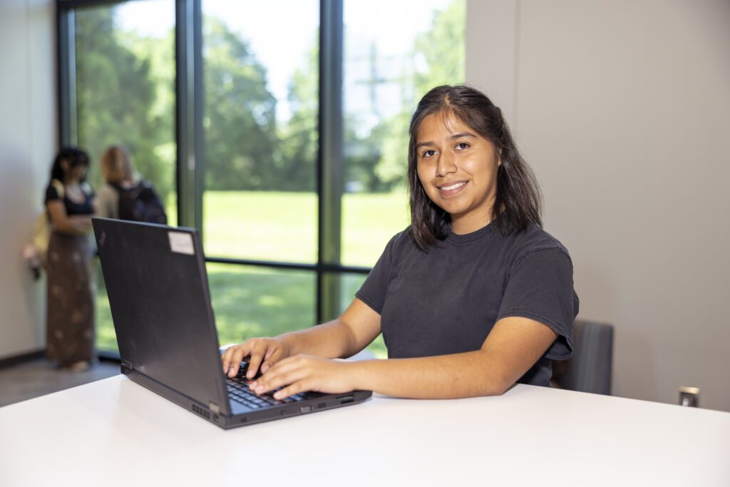 Student on a laptop sitting down by the windows and looking at the camera.