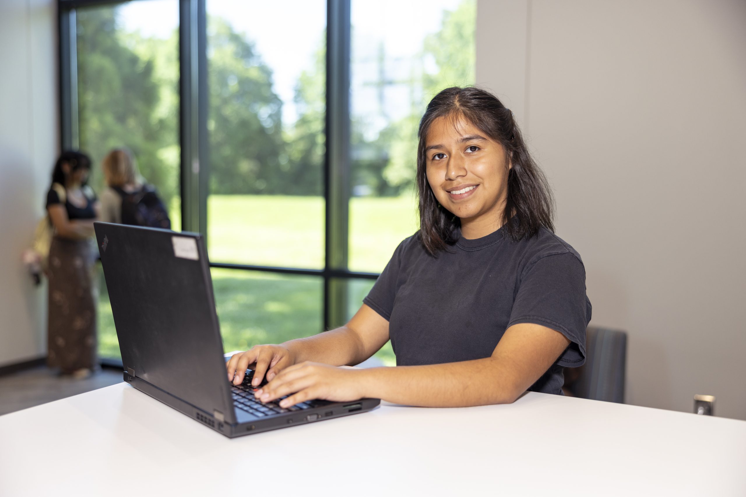 Student on a laptop sitting down by the windows and looking at the camera.