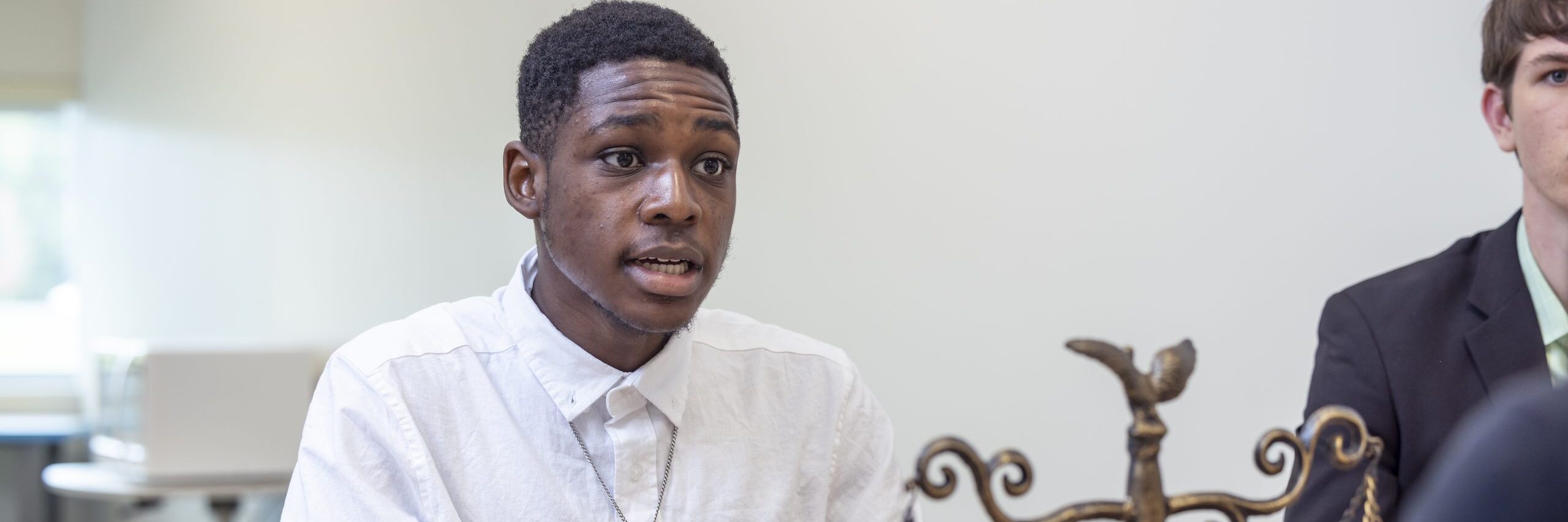 A student dressed in business casual attire seated at a table, attentively looking at a speaker off-camera, with the scale of justice visible nearby.