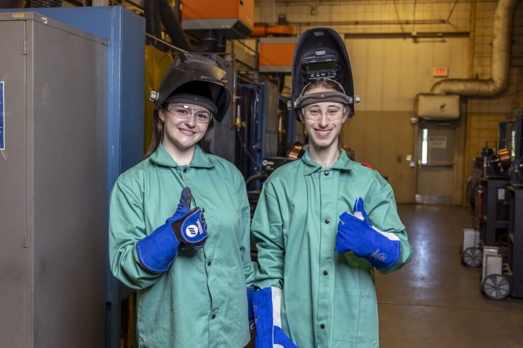 Welding students posing with thumbs up for the camera, their helmets lifted to reveal smiling faces, standing in a workshop setting.