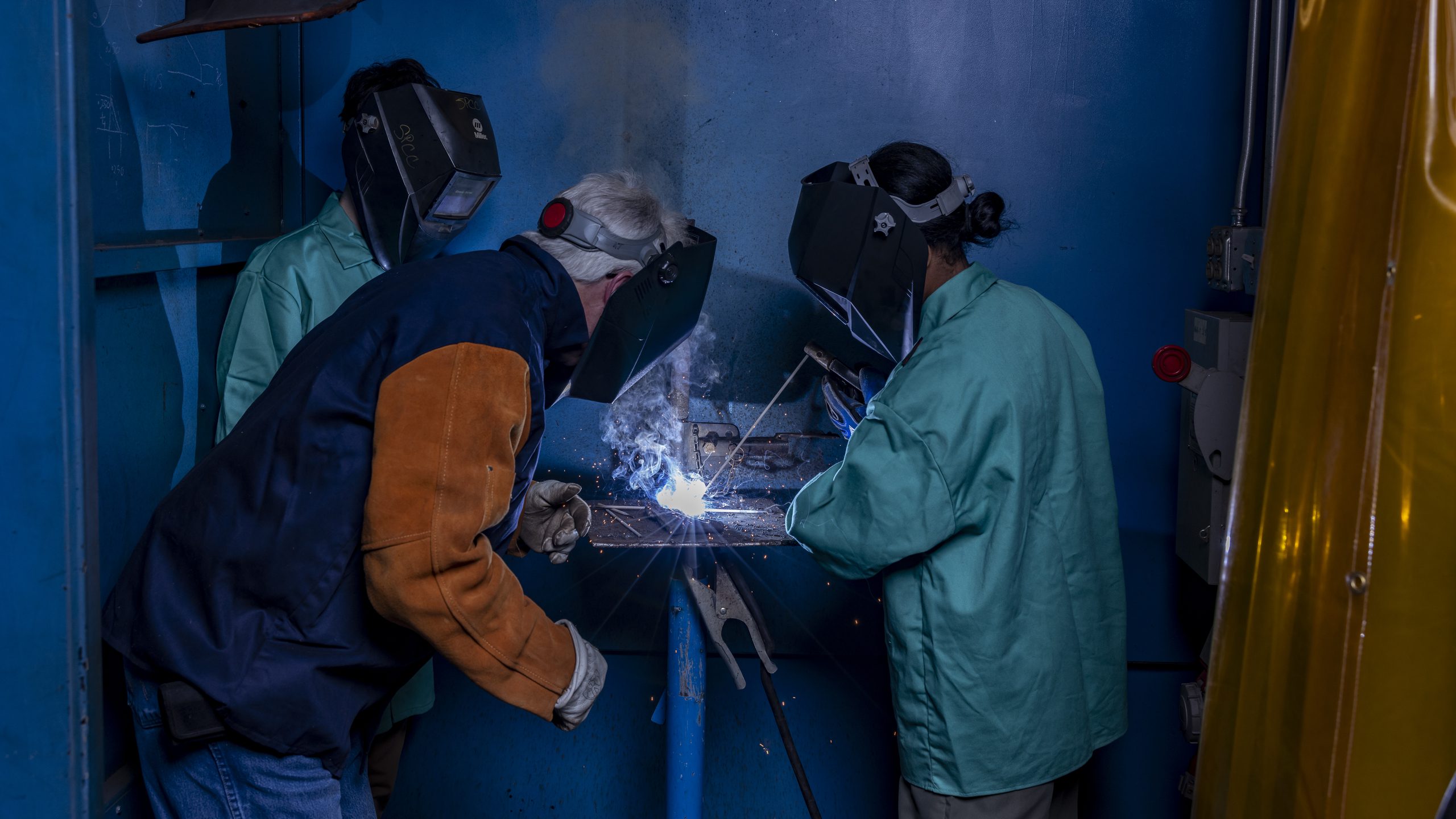 Two students welding while their instructor observes closely, all wearing protective gear in a workshop environment.