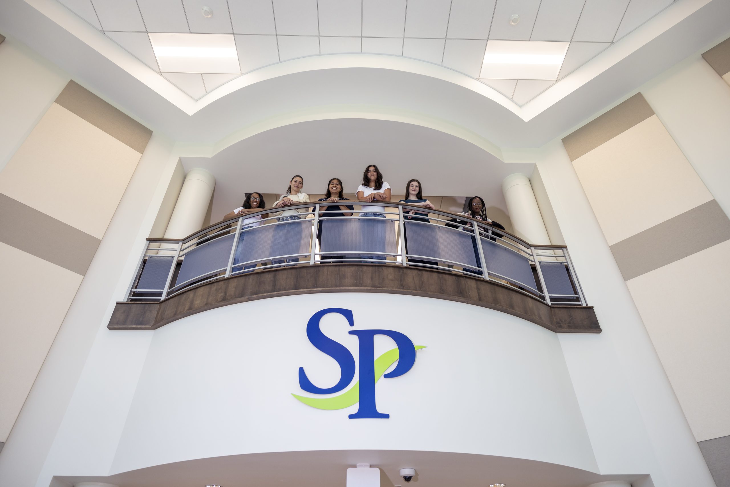 A group of students standing on a balcony, dressed in casual or business casual attire, leaning slightly and looking down.