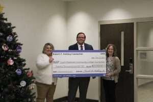 Image of three people holding a large check next to a Christmas tree.