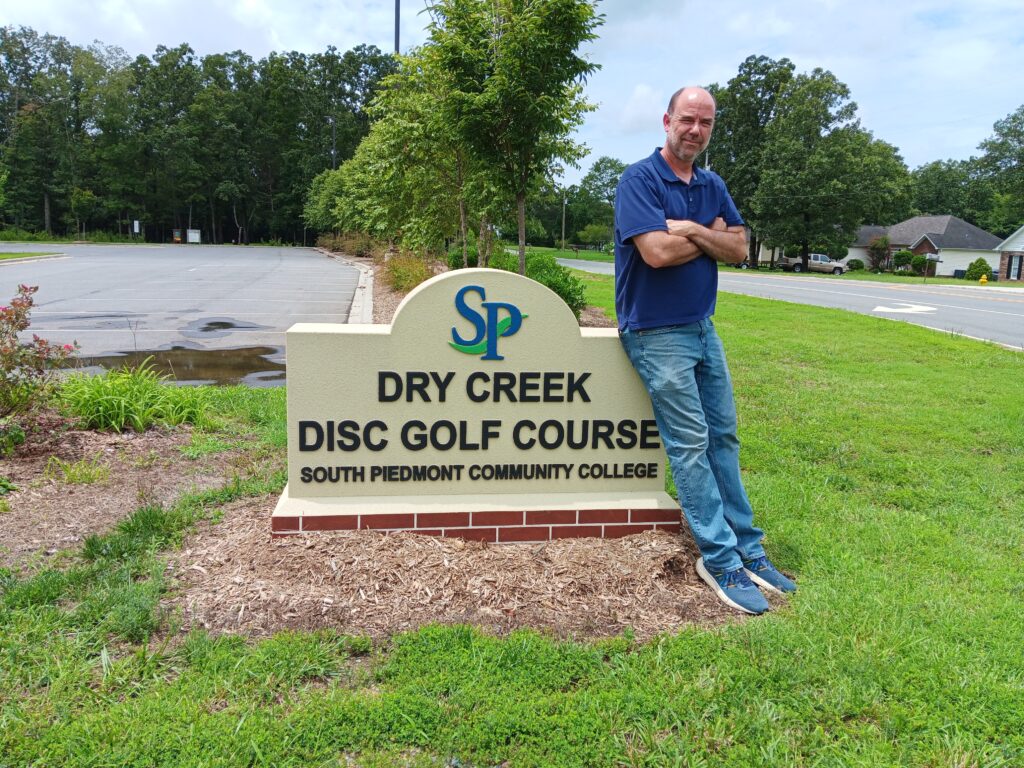 A man standing next to a sign that reads "Dry Creek Disc Golf Course." 