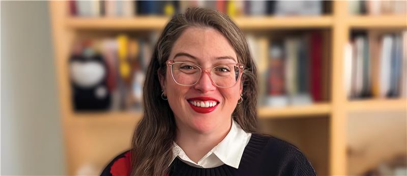 Woman standing in front of a bookcase smiling. She has a black sweater on with red and yellow flowers. She also has red lips and eyeglasses on.