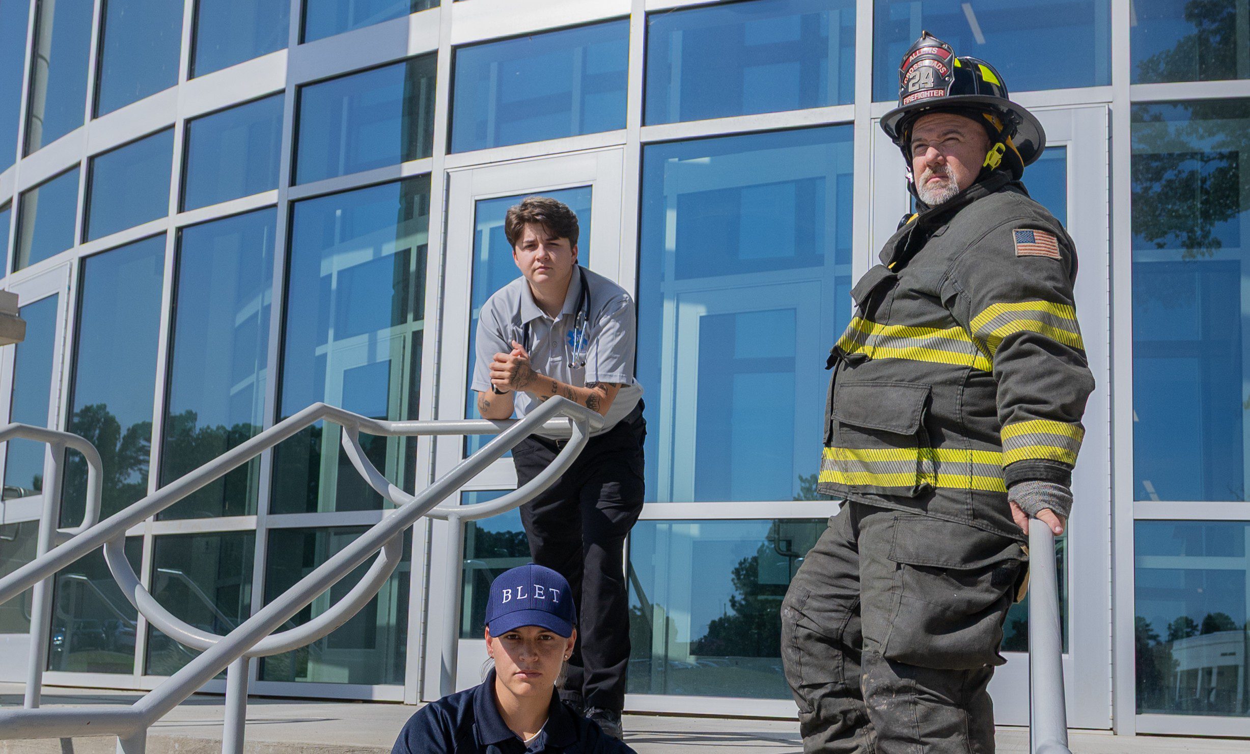 Three Public Safety students at South Piedmont, an EMT student, a Firefighter student, and a BLET student, all strike a serious pose in front of South Piedmont Community College's main building.