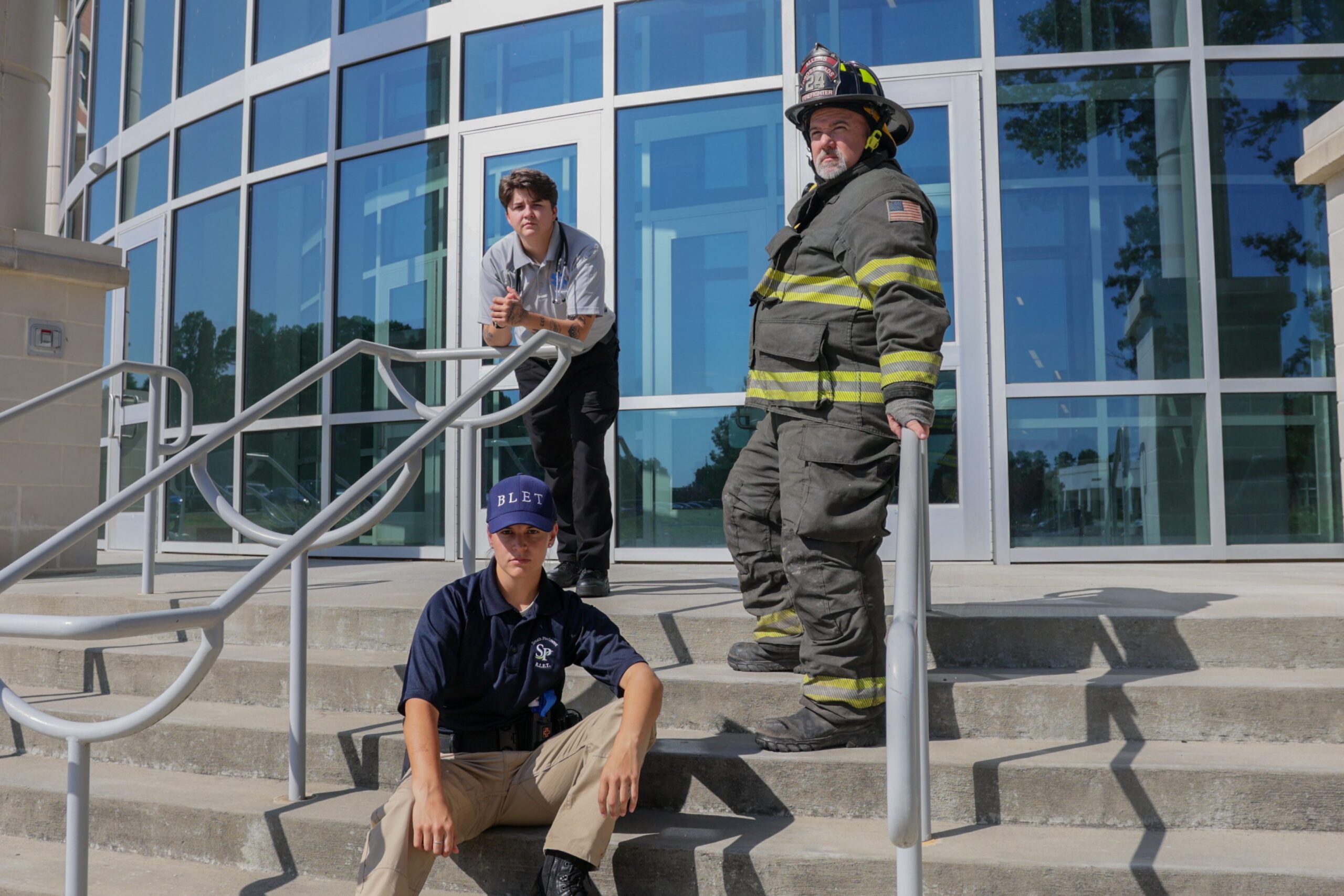 Three Public Safety students at South Piedmont, an EMT student, a Firefighter student, and a BLET student, all strike a serious pose in front of South Piedmont Community College's main building.