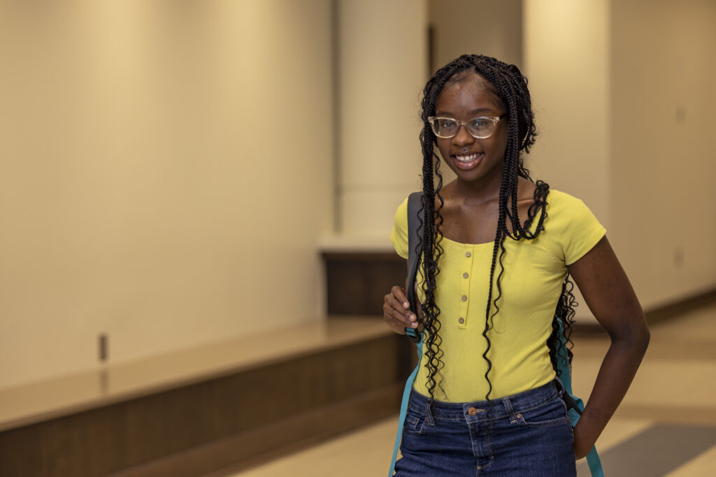 Young female student, dressed in yellow shirt and jeans, stands in a hallway and smiles at the camera.