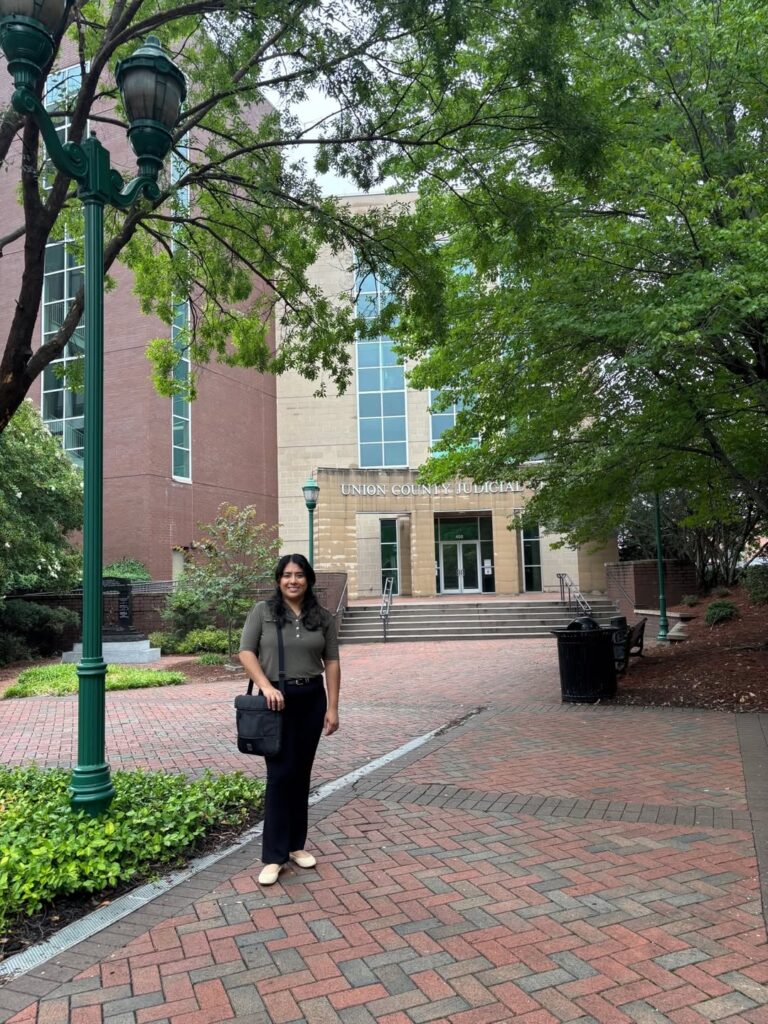 A woman in a grey shirt and black slacks standing under trees outside a courthouse.