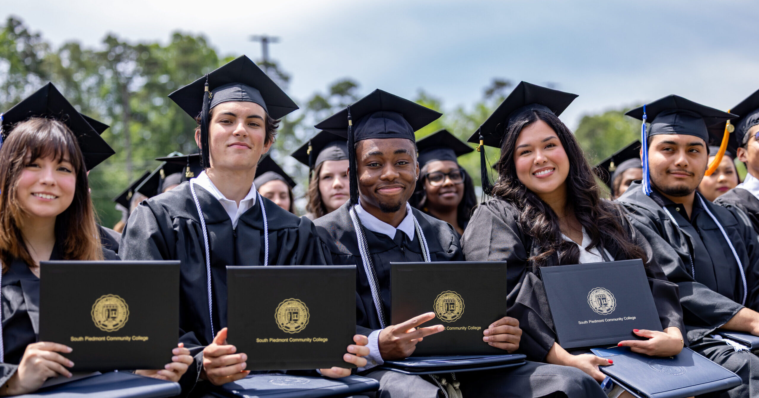 Group of students in their graduation gown, sitting down and smiling at the camera together.