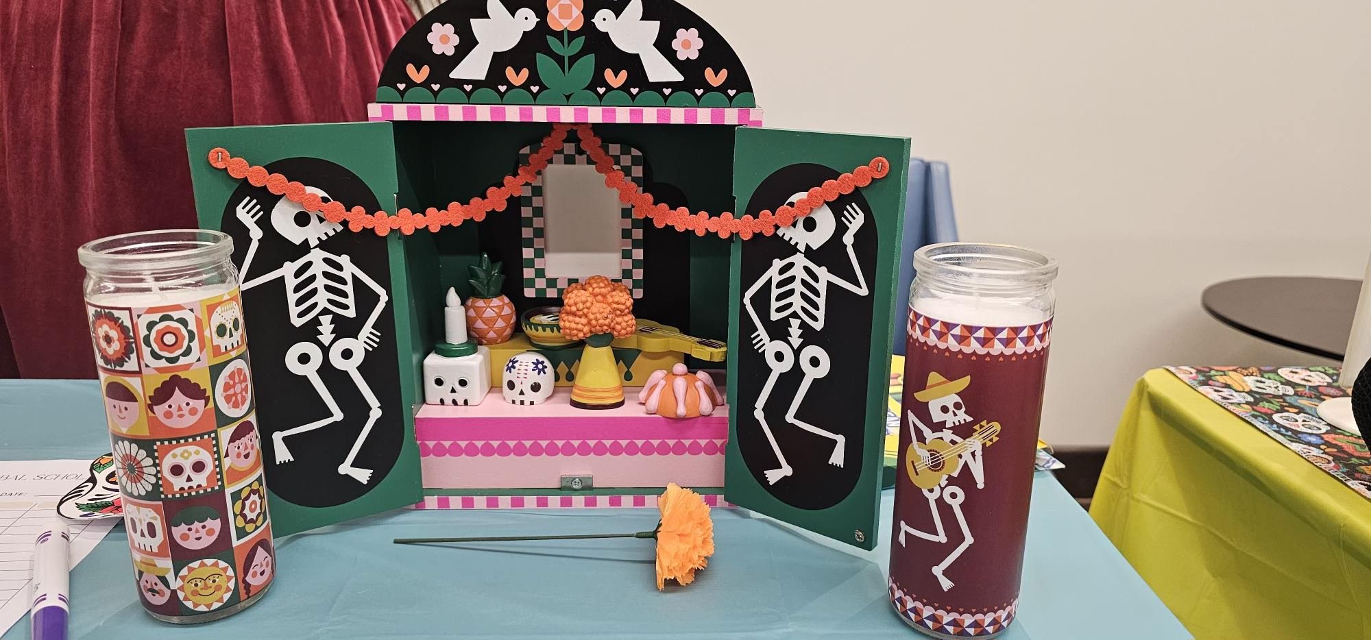 Colorful Día de los Muertos display featuring two decorative sugar-skull candles, dancing skeletons, marigold flowers, and a small altar with traditional decorations.
