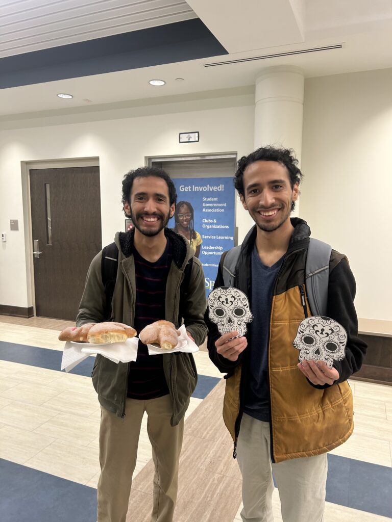 Two smiling people standing indoors at a Día de los Muertos event, holding decorated sugar-skull crafts and pastries.